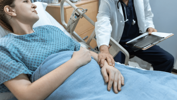 A patient in a blue patterned hospital gown lies in a medical bed. A healthcare professional sits beside her, resting a reassuring hand on the patient's arm while holding a medical file.