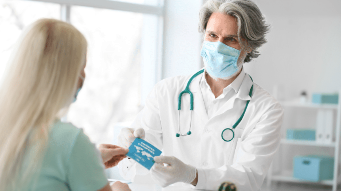A male doctor with grey hair, wearing a face mask and white coat, hands a blue "Immune Passport" document to a blonde female patient.