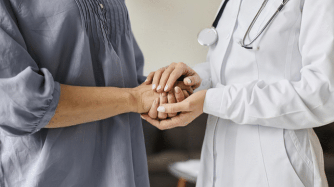 A close-up focused on the hands of a doctor and a patient. The doctor, in a white lab coat, is warmly holding the patient's hands in a gesture of empathy and support.