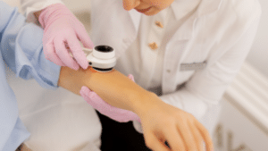 A dermatologist wearing pink gloves uses a dermatoscope to examine a mole on a patient’s forearm. The doctor is wearing a white lab coat and is leaned over the patient's arm to get a clear view through the magnified lens.