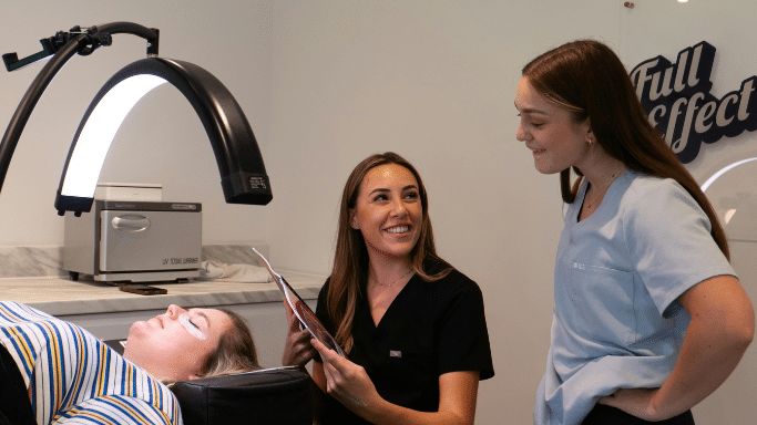 Two female healthcare professionals in scrubs are smiling and talking in a clinical setting. One holds a clipboard. A patient lies on a treatment bed in the foreground under a curved, professional LED light.