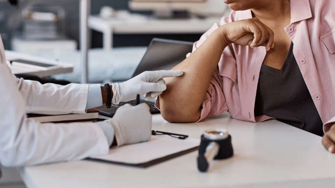 A doctor in a white coat and gloves points to a spot on a patient's elbow. A dermatoscope sits on the white desk between them, and the doctor appears to be taking notes on a clipboard.
