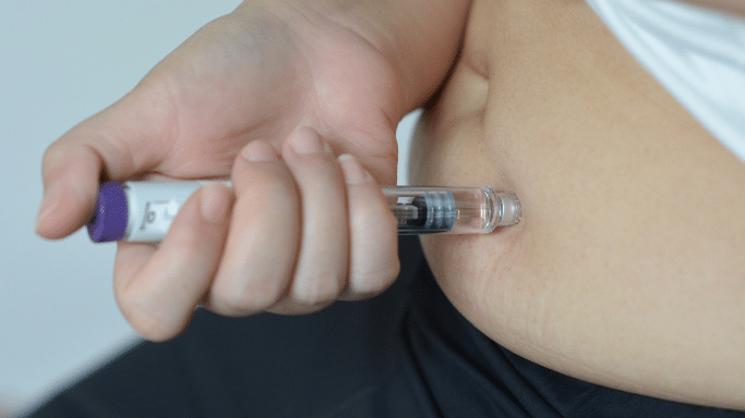 Close-up of a person's hand administering a subcutaneous injection into their abdomen using a clear plastic medical pen with a purple end cap.