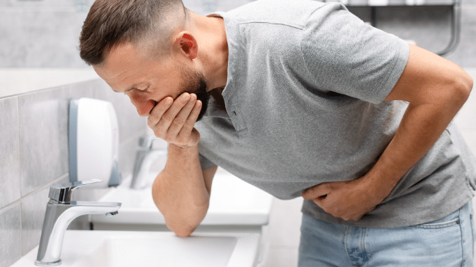 A medium shot of a man with a beard leaning over a white sink in a bathroom, holding one hand over his mouth and the other over his stomach with a pained expression, indicating nausea or sickness.