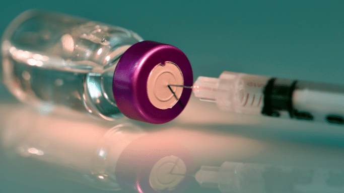 Extreme close-up of a medical syringe needle piercing the rubber stopper of a glass vial with a purple cap. The scene has a reflective surface beneath and a muted teal background.
