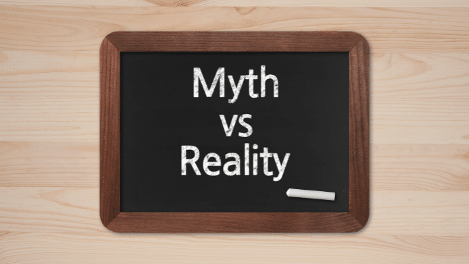 A small wooden-framed blackboard sitting on a light-colored wood surface. Written on the chalkboard in white text is "Myth vs Reality," with a single piece of white chalk resting at the bottom right.
