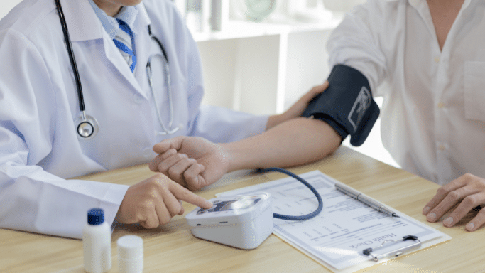 A doctor in a white lab coat and stethoscope places a blood pressure cuff on a patient's arm and presses a button on a digital monitor sitting on a wooden desk alongside health forms.