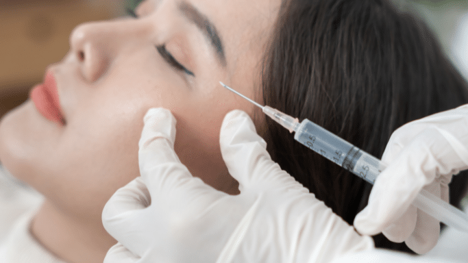 An angled close-up shot of a person resting horizontally during a cosmetic procedure. A practitioner in white latex gloves uses one hand to gently stretch the skin near the lower eyelid and cheekbone while aiming a clear fluid-filled syringe toward the area.
