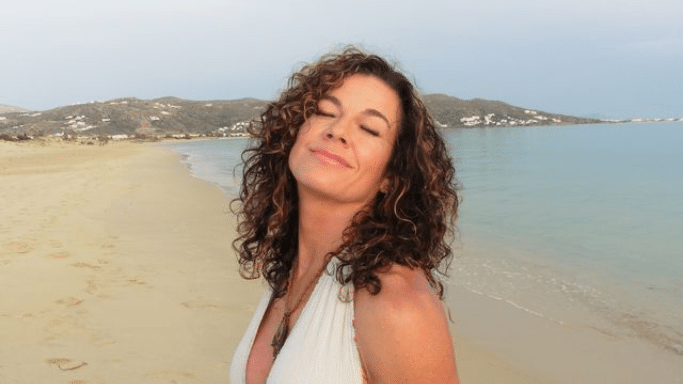 A woman with curly brown hair smiling with her eyes closed on a sandy beach next to the ocean.