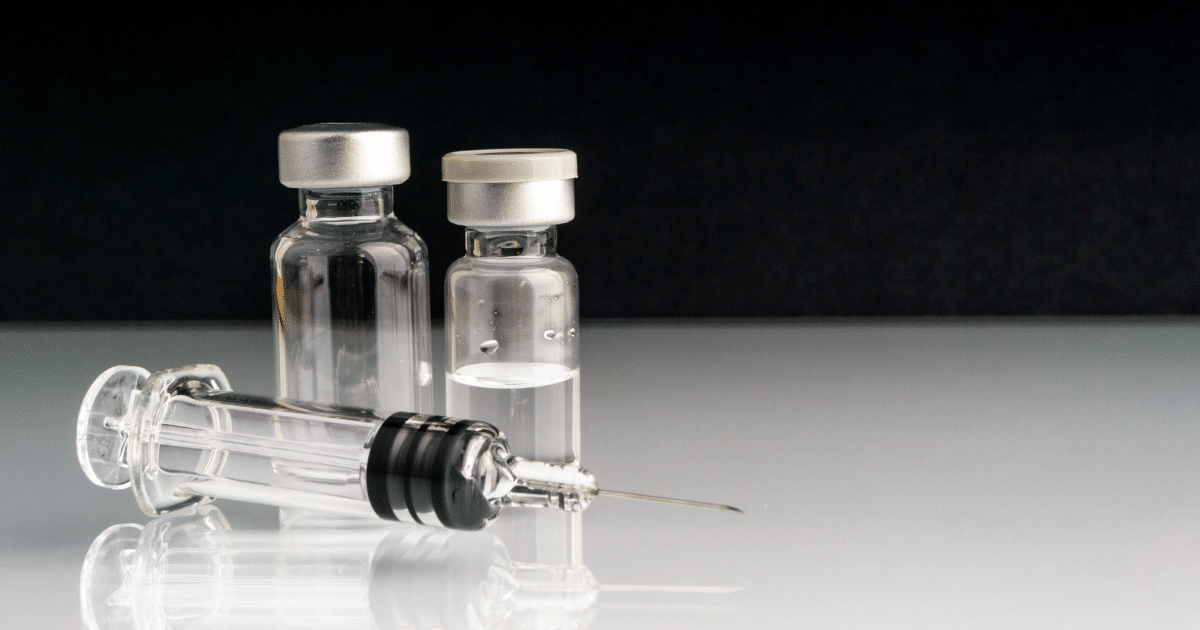 A glass medical syringe lies horizontally in the foreground on a reflective surface, with its needle pointing to the right. Behind it stand two small, clear glass medicine vials with metallic caps, set against a solid dark gray background.