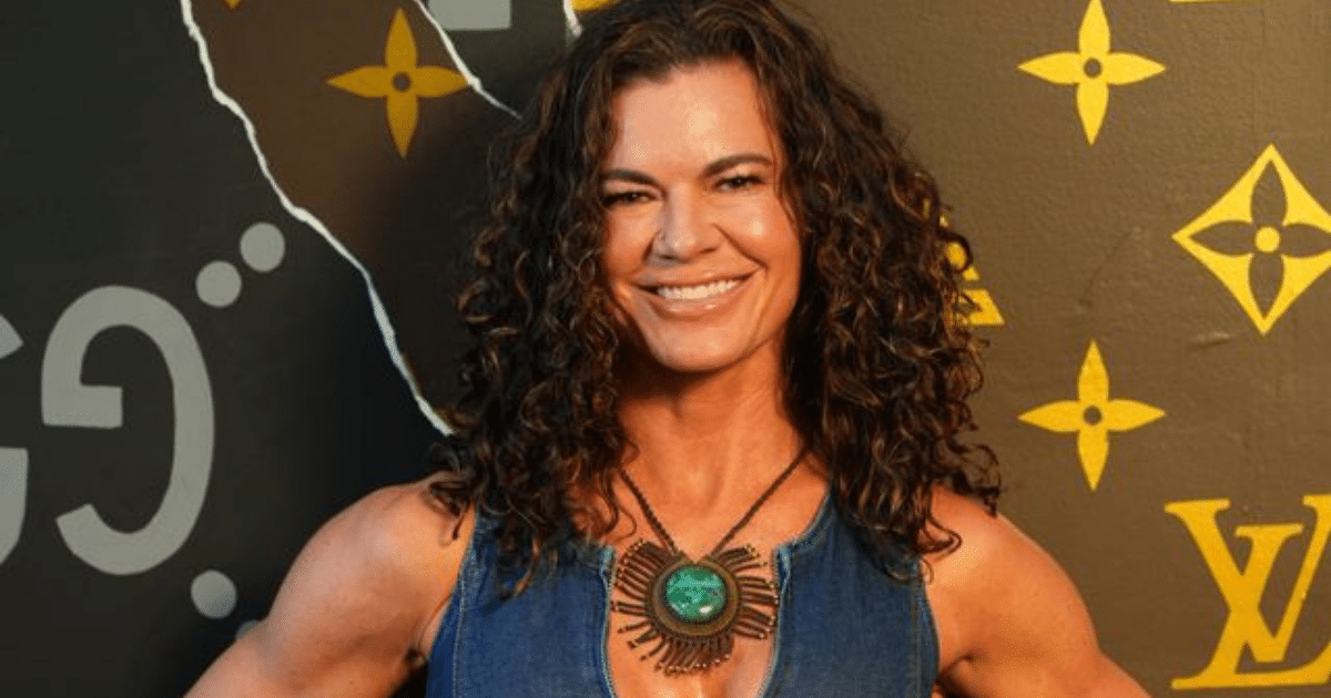 A smiling woman with voluminous, shoulder-length curly brown hair. She is wearing a blue denim top and a large, detailed circular statement necklace with a green stone center.