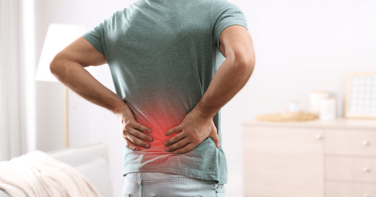 A medium shot from behind of a person in a green t-shirt and jeans holding their lower back with both hands, featuring a glowing red overlay on the lower spine area to represent back pain.