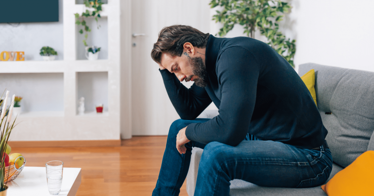 Side view of a bearded man in a black turtleneck and jeans sitting on a living room sofa, leaning forward with his head in his hand, looking downcast or stressed.