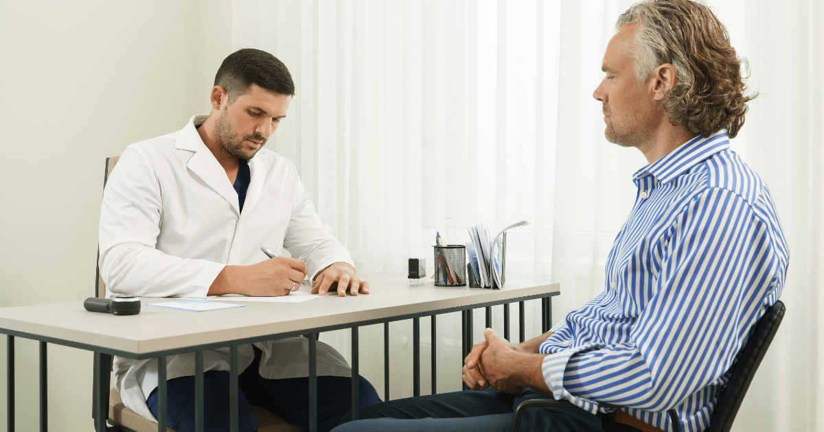 A male doctor in a white lab coat sits at a light-colored desk, looking down and writing on a piece of paper with a pen. Across from him sits a middle-aged male patient with wavy gray hair, wearing a blue and white striped button-down shirt. The patient is sitting in profile, looking towards the doctor with an attentive expression.