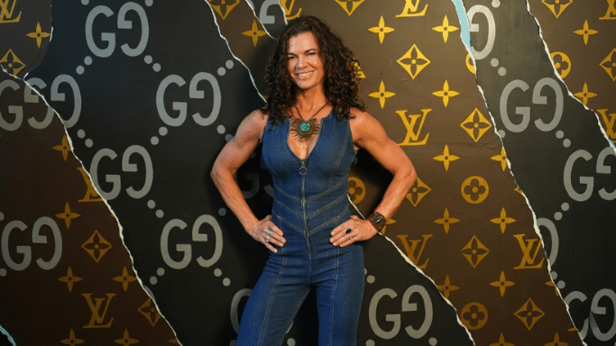 A smiling woman with long, curly dark hair stands with her hands on her hips. She is wearing a blue denim sleeveless jumpsuit with a front zipper and a large statement necklace. The background features a "torn paper" effect, revealing repeating Gucci and Louis Vuitton luxury logos.