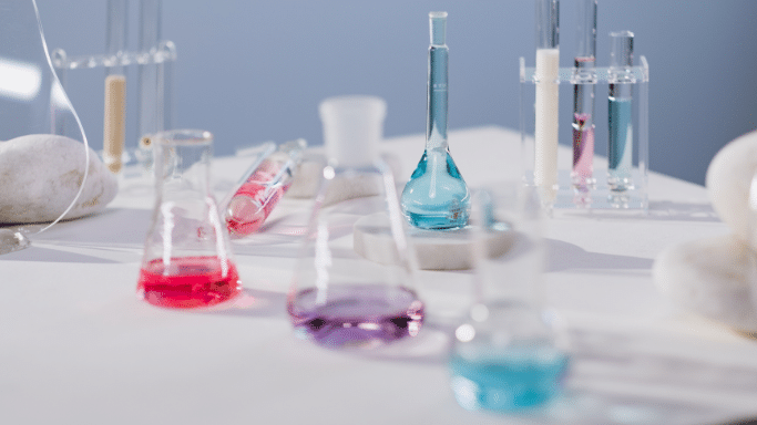 A bright, clean laboratory setting featuring various glass flasks and test tubes on a white table. The containers are filled with liquids in vibrant shades of pink, purple, and blue. Smooth white stones are scattered around the table, adding a serene, aesthetic feel.