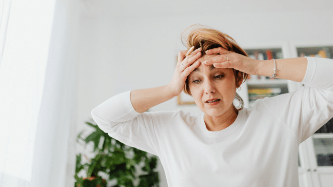 A middle-aged woman with short blonde hair looks visibly distressed or overwhelmed. She has both hands pressed against her forehead and her eyes are directed downward with a pained expression. She is wearing a simple white long-sleeved shirt in a brightly lit room.