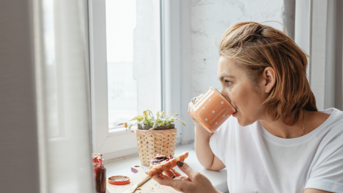 A profile view of a woman drinking from an orange polka-dot mug while looking out a bright window. In her other hand, she holds a piece of toast topped with red jam. A small potted plant and a jar of jam sit on the windowsill beside her.