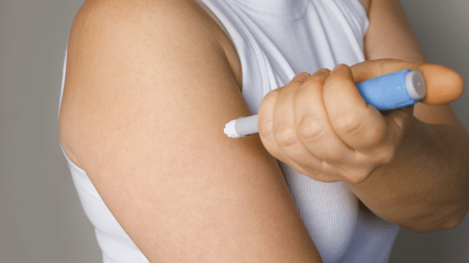 A close-up shot of a person’s upper arm as they use a blue and grey insulin pen to administer an injection. The person is wearing a white sleeveless top, and the focus is on the precision of the medical device against the skin.