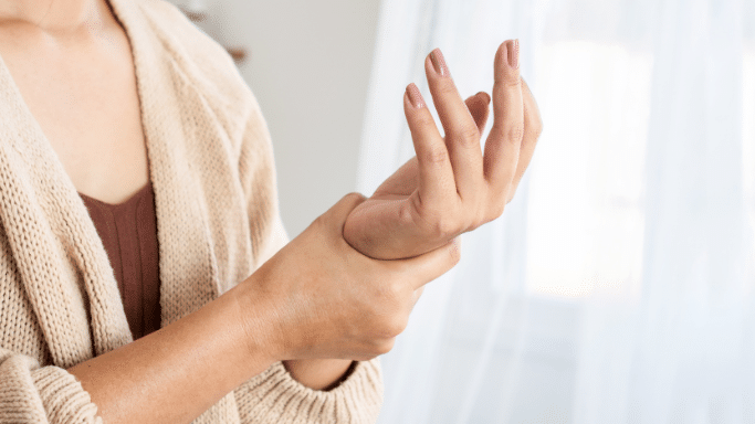 A close-up of a woman’s hands as she rubs her left wrist with her right hand, suggesting pain or discomfort. She is wearing a beige knit cardigan over a brown top. The background is soft-focus, showing a bright window with white curtains.
