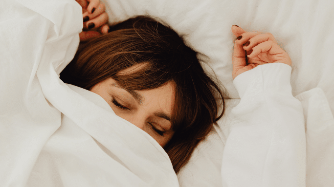 A close-up, top-down shot of a woman with brown hair sleeping peacefully in a bed. Her eyes are closed, and her lower face is tucked beneath a white blanket. She is wearing a white sweatshirt, and her hands, featuring dark nail polish, rest gently on either side of her head against the white pillow.