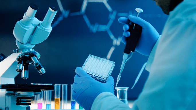 A scientist wearing blue nitrile gloves uses an electronic pipette to dispense liquid into a clear 96-well microplate. A modern microscope sits to the left, and vibrant test tubes containing colored solutions are lined up in front of a dark background displaying chemical structures.