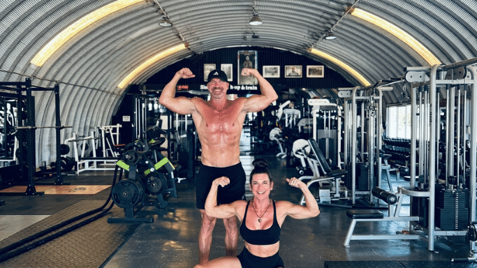 A shirtless, muscular man with a black baseball cap and a muscular woman in a dark sports bra and shorts strike a double-bicep pose together inside a large, hangar-style gym with a curved corrugated metal roof. A wide variety of weightlifting equipment fills the background.
