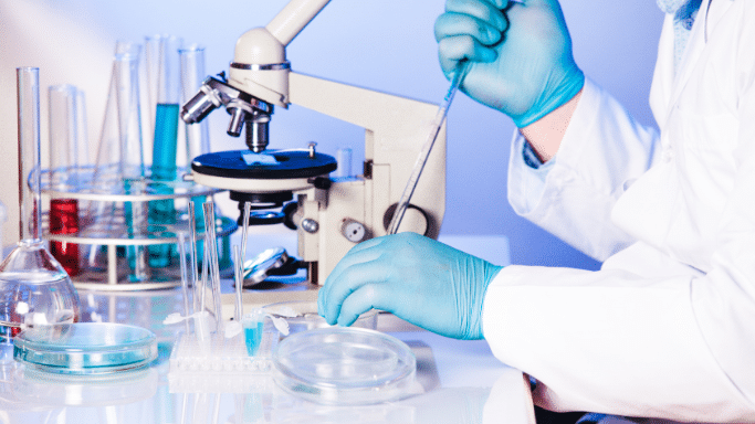 A bright laboratory setting showing a researcher in a white lab coat and blue gloves. One hand rests on a petri dish near a microscope, while the other hand uses a glass pipette. Multiple test tubes filled with colorful liquids are visible in a rack on the left side of the frame.
