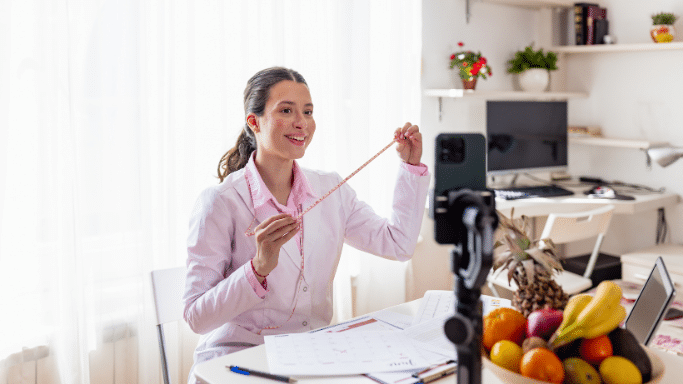 A smiling female dietitian or medical professional wearing a white coat sits at a desk, holding up a pink measuring tape toward a smartphone mounted on a tripod for a video recording. The desk has calendars, papers, and a large bowl of fresh fruit, including bananas, oranges, and a pineapple.