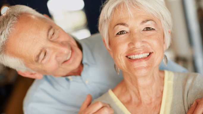 A close-up portrait of a happy senior couple. An older woman with short white hair smiles warmly directly at the camera, while her husband stands closely behind her, looking down at her with an affectionate smile.