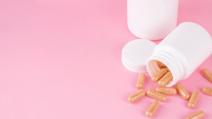 Two white plastic supplement bottles on a solid pink background. One bottle is upright, while the other is overturned, spilling out several brown, clear-shelled herbal capsules.