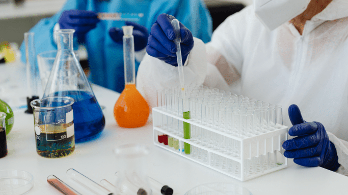 A researcher wearing a white hazmat suit and dark blue nitrile gloves uses a pipette to drop liquid into a rack of glass test tubes. Various laboratory flasks filled with blue and orange fluids sit on the white table.