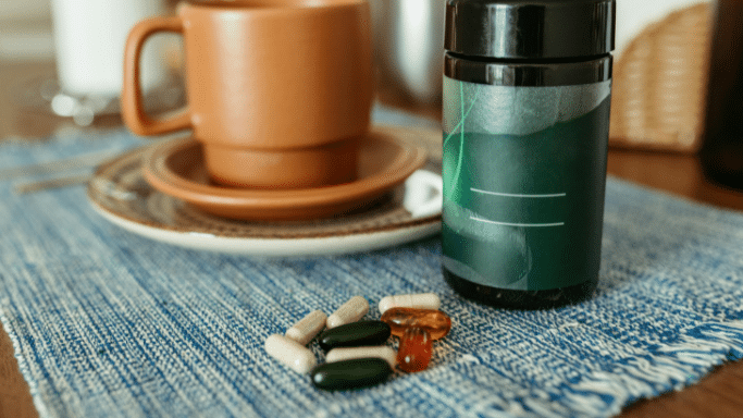 A variety of loose vitamins and supplements—including green softgels, amber gel caps, and tan capsules—are scattered on a blue woven placemat next to a dark green bottle. A brown ceramic coffee mug sits on a saucer in the background.