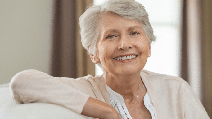 A warm portrait of a senior woman with short, styled white hair and elegant earrings. She is wearing a cream-colored cardigan and smiling brightly while leaning against a white sofa cushion.