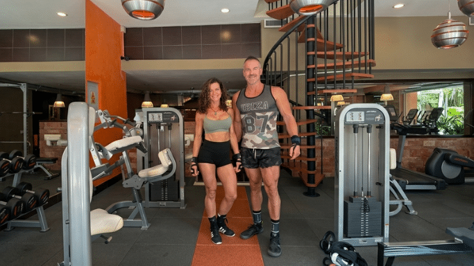 A fit woman in a light green sports bra and black athletic shorts stands beside a tall, muscular man wearing a camouflage tank top with "IBIZA 87" printed on it and dark shorts. They are posing together on an orange floor runner inside a commercial fitness center, with a prominent wooden spiral staircase and various workout machines in the background.
