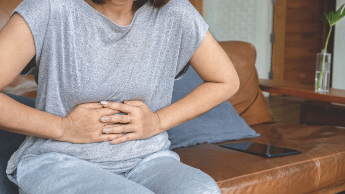 A close-up shot of a woman sitting on a brown leather couch, dressed in a casual gray t-shirt and light gray sweatpants. She is clutching her stomach with both hands, showing signs of physical discomfort or abdominal pain.