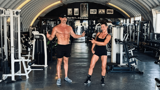 An athletic, shirtless man in a black baseball cap and dark shorts stands next to a muscular woman in a black crop top and black shorts. They are inside a large, hangar-style gym featuring a curved corrugated metal roof. The man gives a thumbs-up while the woman smiles brightly and points over at him.
