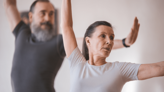 A senior couple intently practicing a yoga or stretching pose together. In the sharp foreground, an older woman with dark hair pulled back extends her arms smoothly with a focused expression. Behind her and slightly out of focus, a senior man with a full gray beard mirrors her arm movements.