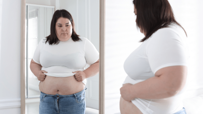 A woman stands in front of a mirror in a bright room, lifting her white t-shirt to look at her stomach in her reflection. She is wearing blue jeans and has a focused, neutral expression.