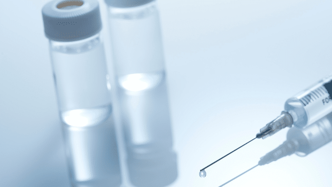 A close-up, high-angle shot of a medical syringe lying on a reflective surface. A single clear drop of liquid hangs from the needle tip, with two blurry glass vials in the background.