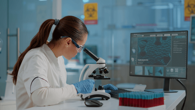 A scientist in a white lab coat, safety glasses, and blue gloves works in a laboratory. She is looking through a microscope while her left hand rests on a computer keyboard. A monitor in the background displays a cellular image.