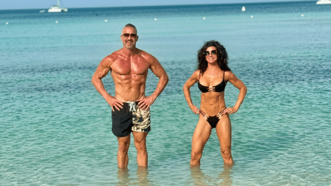 A muscular man and woman stand waist-deep in clear turquoise ocean water. Both are tanned, wearing swimwear and sunglasses, and posing with their hands on their hips against a bright beach background.