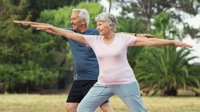 An elderly man and woman are practicing a yoga or stretching pose outdoors in a park. They are both smiling and extending one arm forward while lunging, with green trees in the background.