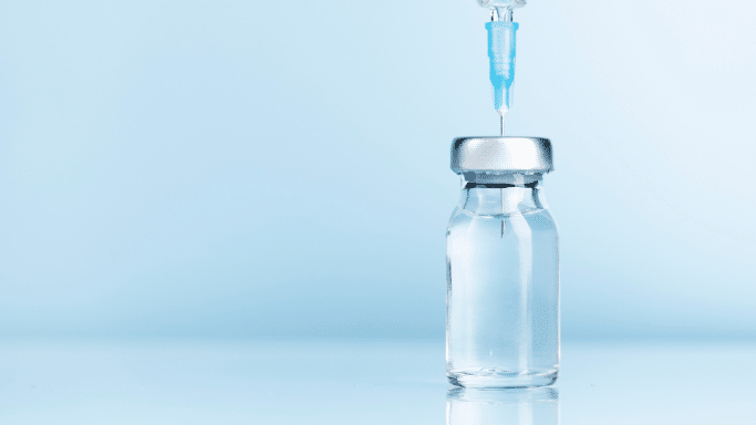 A close-up shot of a small, clear glass medical vial filled with liquid, sitting against a solid, light blue background. A syringe with a blue needle hub is inserted straight down through the rubber stopper of the vial, preparing to draw out medication.