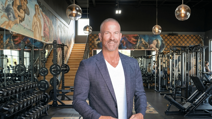 A middle-aged man with a shaved head and a smile stands in the middle of a modern gym. He is wearing a dark gray blazer over a casual white v-neck t-shirt. The background features gym equipment, racks of dumbbells, a wooden staircase, and large wall murals depicting classical art.