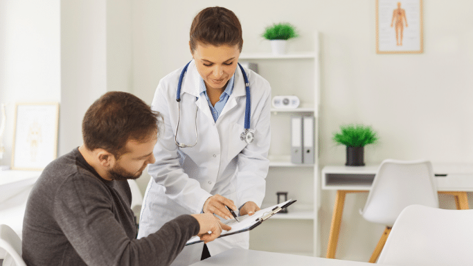 In a bright medical office, a female doctor in a white coat with a stethoscope over her shoulders stands next to a seated male patient. She holds a clipboard with medical documents, pointing down at a section with a pen as she explains it to him. The patient looks closely at where she is pointing.