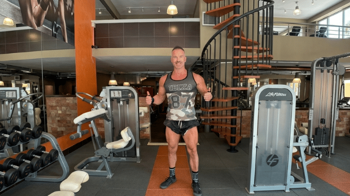 A muscular man stands in the center of a spacious gym floor, giving a double thumbs-up to the camera. He is wearing a camouflage tank top with the number 87 on it, black athletic shorts, and sneakers. The gym behind him includes weight machines, a spiral staircase, and an upper mezzanine level.