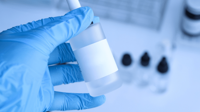 A close-up view of a hand wearing a bright blue nitrile glove holding a small, frosted glass dropper bottle with a blank white label. The background is a clean, slightly out-of-focus laboratory or medical setting with other small bottles visible.