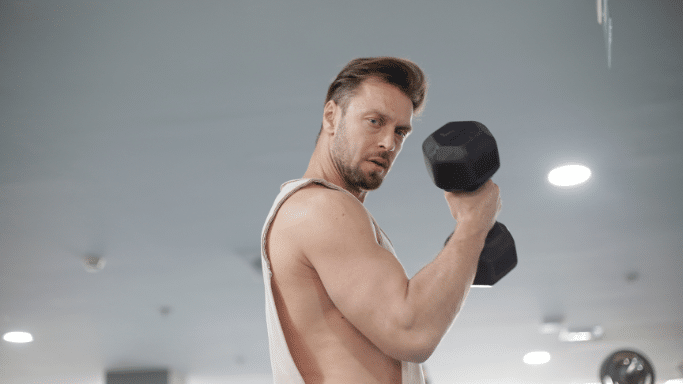 A close-up, low-angle shot of a fit man in a gym performing a bicep curl with a black hexagon dumbbell. He is wearing a white tank top, has short brown hair, and looks intensely toward the camera as he lifts the weight.