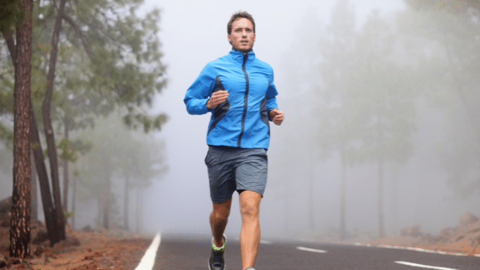 A man in a blue athletic jacket and gray shorts is running down the center of an empty asphalt road. The background is filled with tall pine trees shrouded in heavy, low-lying white fog, creating a misty atmosphere.